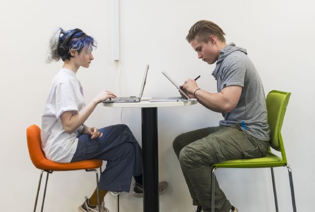 A male student and a female student, sitting opposite each other on a raise table, both working on laptops