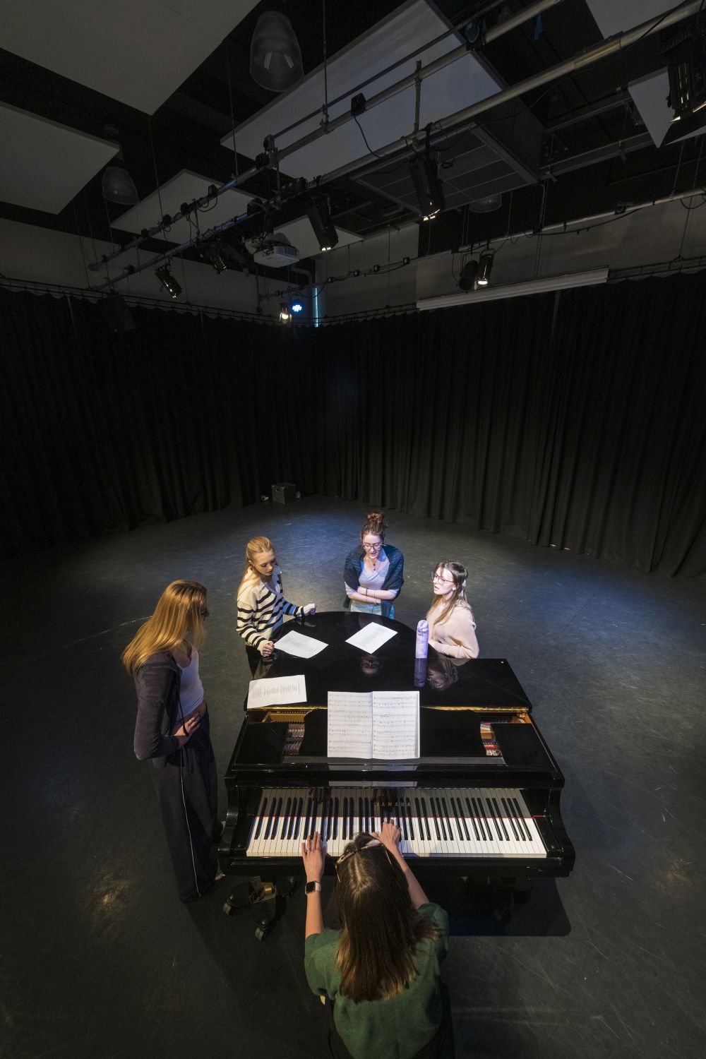 A group of female students singing gathered around a  large piano in a  black theatre